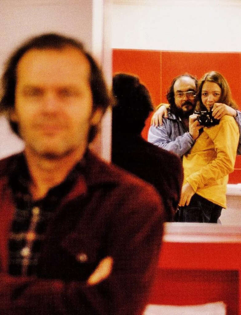 A blurred man in the foreground faces the camera, while in the background, a man in glasses embraces a woman holding a camera, both reflected in a mirror. The room features red and white walls.