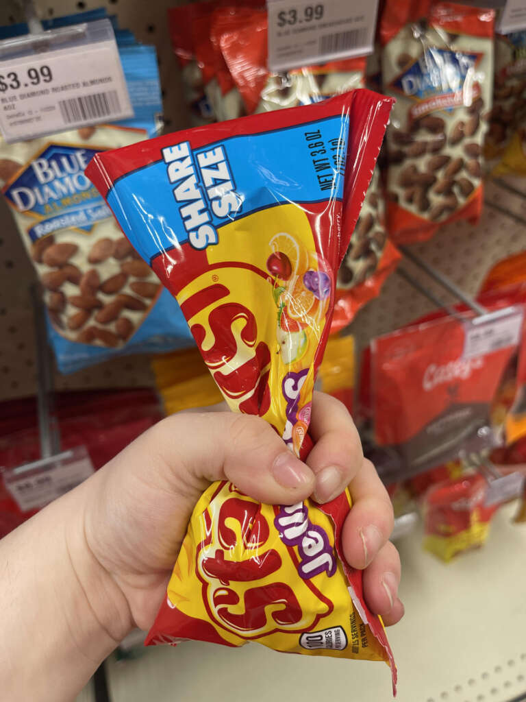 A hand holding a share-size bag of Skittles at a store. Shelves in the background display various snacks, including Blue Diamond almonds and candy. Price tags are visible on the shelf edges.