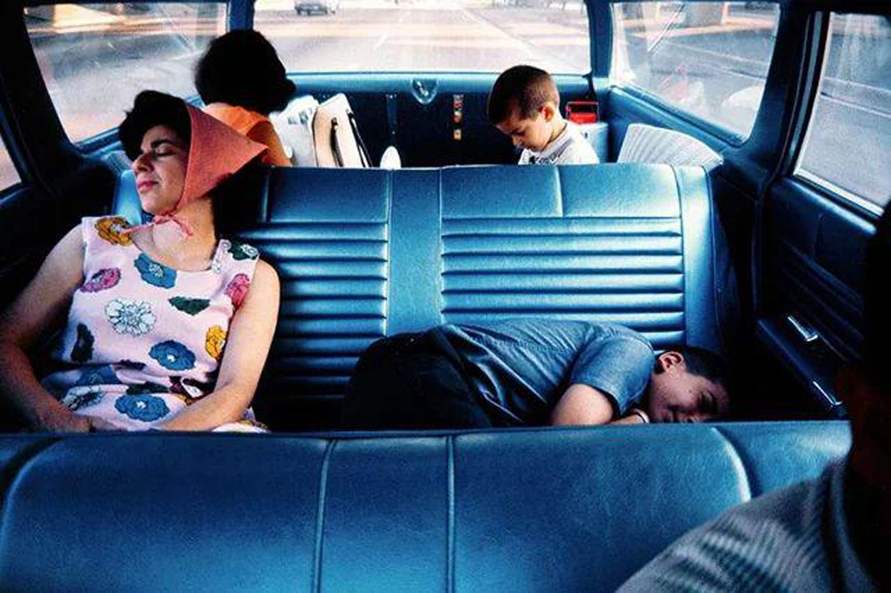 A woman and three children are asleep in a vintage car with blue leather seats. The woman is wearing a floral dress and pink headscarf, while two children rest against the seats and one is lying down. The view outside shows a road.