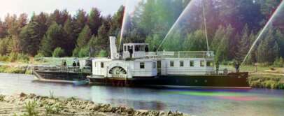 A vintage riverboat travels along a calm river, with trees in the background. The side features a large paddle wheel, and water sprays upward from spigots on the deck. The scene has a colorful, slightly faded quality.