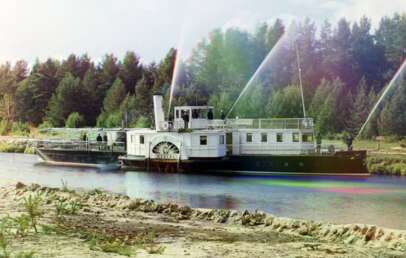 A vintage riverboat travels along a calm river, with trees in the background. The side features a large paddle wheel, and water sprays upward from spigots on the deck. The scene has a colorful, slightly faded quality.