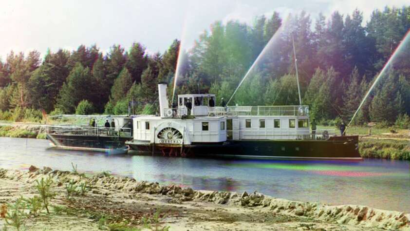 A vintage riverboat travels along a calm river, with trees in the background. The side features a large paddle wheel, and water sprays upward from spigots on the deck. The scene has a colorful, slightly faded quality.