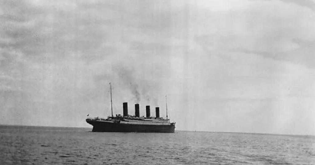 A black and white photo of a large ocean liner with four smokestacks sailing on a calm sea. Smoke is billowing from the stacks into a cloudy sky. The ship is centered and moving from left to right.