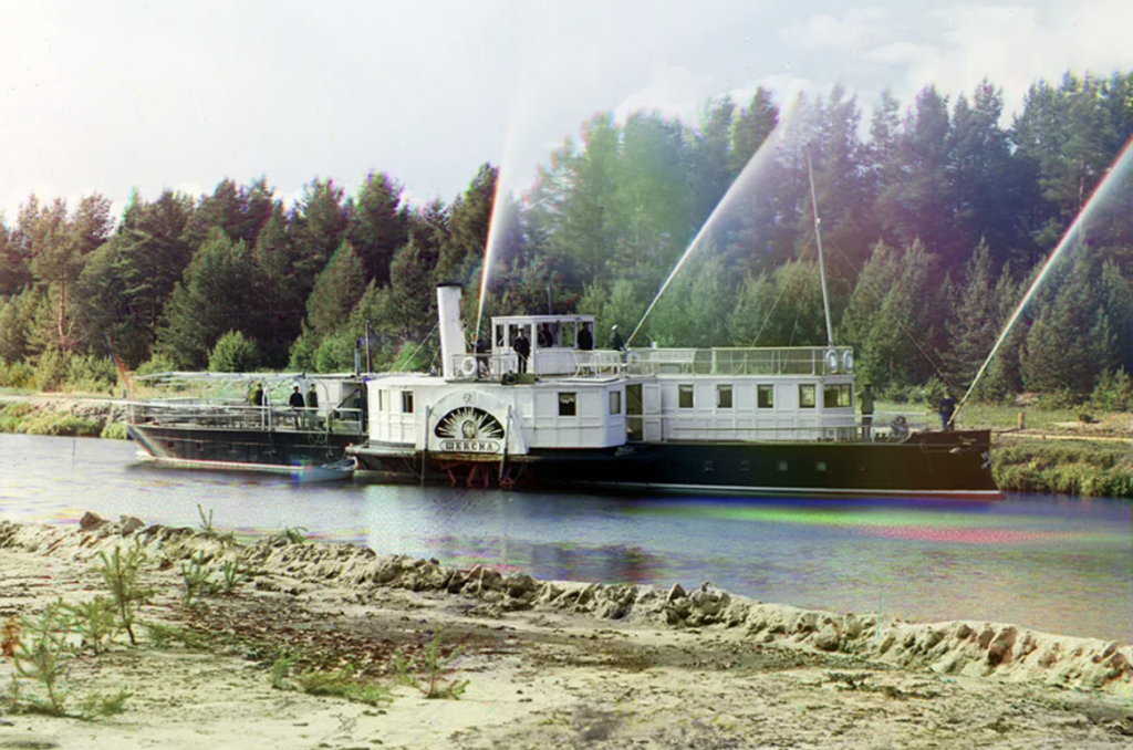 A vintage steamboat travels along a narrow river surrounded by dense forest. The boat has a paddlewheel and emits trails of smoke from its chimney. The clear sky and calm water reflect a serene and historical setting.