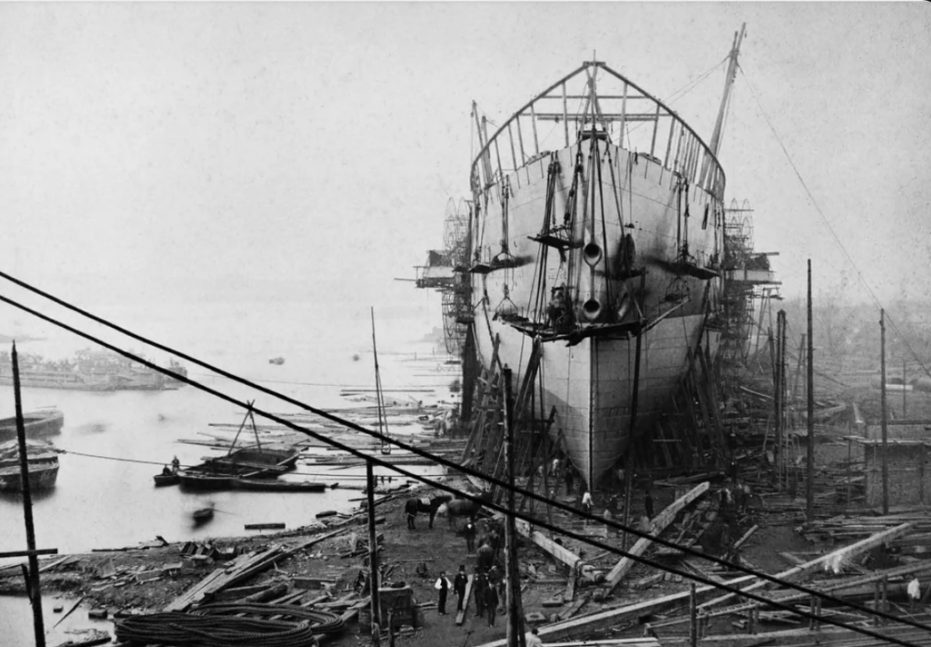 A large ship under construction is on a dry dock near a waterfront. Workers and scaffolding surround the vessel. Several boats float in the water, and numerous construction materials are scattered on the ground. The sky is overcast.