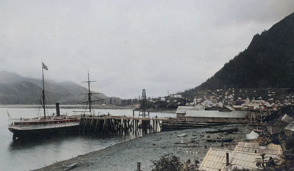 A vintage photograph of a coastal village with a large ship docked at a wooden pier. The foreground shows a rocky shore, while the background is filled with dense forested hills. Small buildings are scattered along the shoreline.