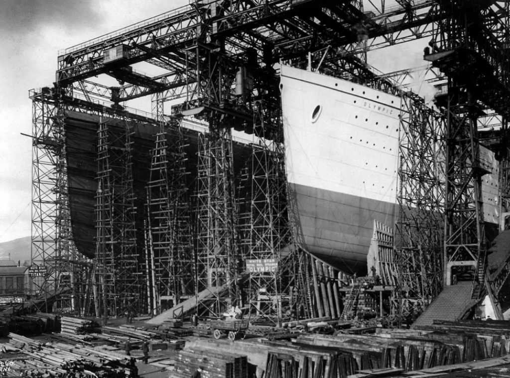 The image shows a large ship under construction in a shipyard. The hull is partly painted and elevated by a scaffold. Cranes and scaffolding surround it, and workers are seen on the ground, among construction materials.