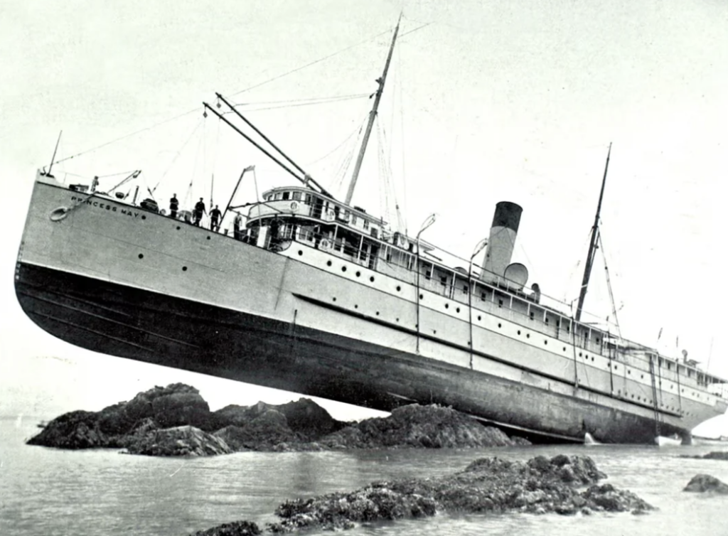 A vintage photograph of a large steamship stranded on rocks. The ship is tilted and elevated above the water, with several people visible on deck. The image is black and white, emphasizing the ship's dramatic position.