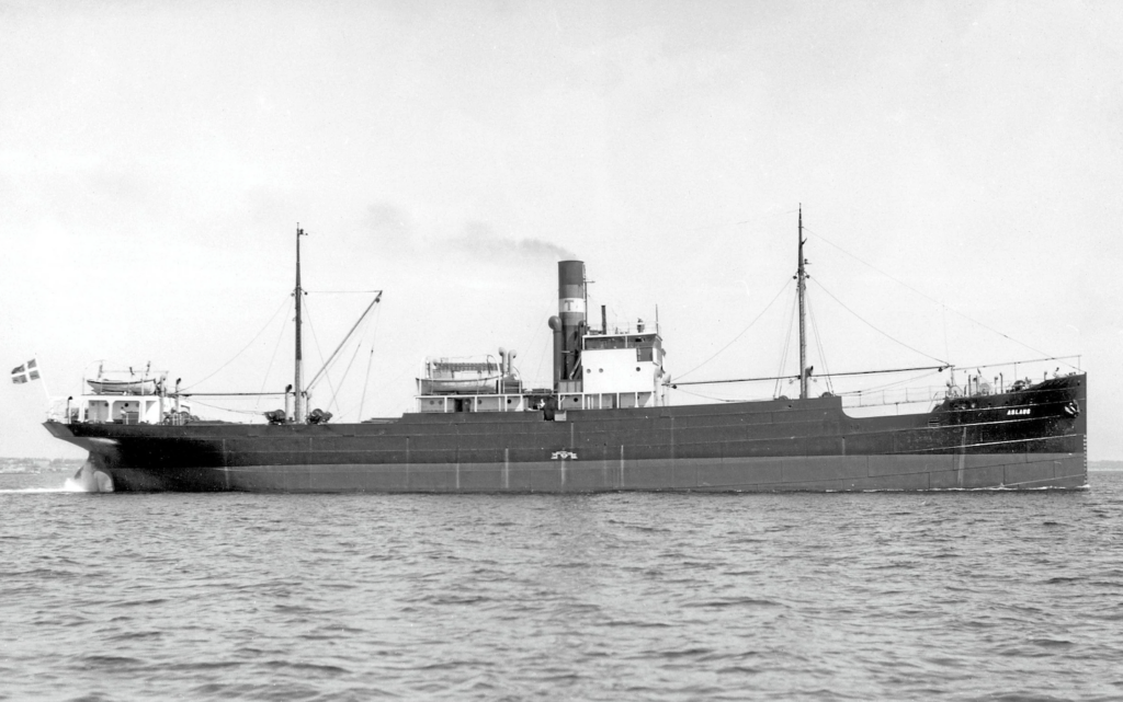 Black and white photo of a large cargo ship at sea. The vessel has a central smokestack and two tall masts; the flag at the stern is visible. The hull and superstructure are prominent against the water and sky.