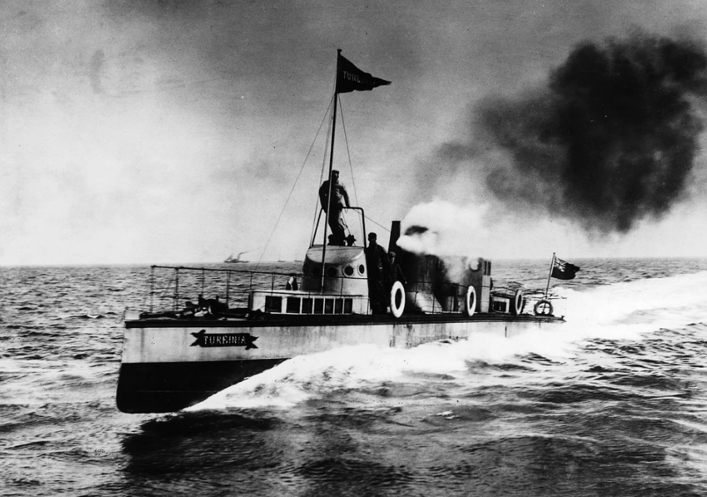 A black and white photo of the Turbinia, an early steam-powered ship, moving at high speed on water. Smoke billows from its funnel, and several crew members are visible on deck. The sea appears choppy under a cloudy sky.