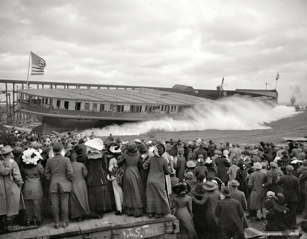 A large crowd watches the dramatic launch of a ship in a shipyard. Water splashes as the vessel slides into the water. The scene is from an earlier era, with people dressed in vintage clothing and hats. An American flag is visible on the ship.