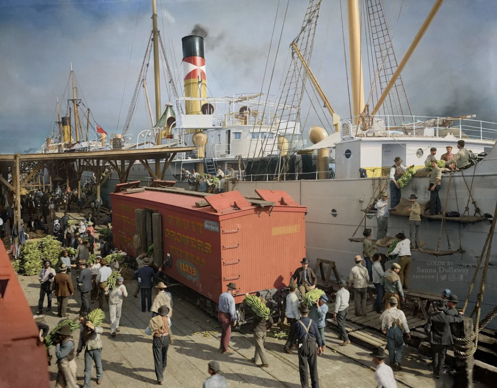 A bustling port scene with workers loading a large cargo ship and a railroad boxcar. People carry bunches of bananas, and the ship has a tall smokestack with colorful markings. The sky is clear, indicating a busy, productive day at the dock.