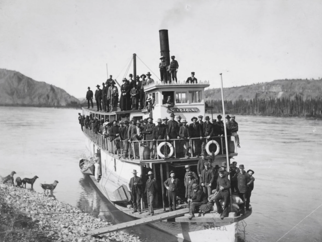 Black and white photo of a riverboat named "Nora" crowded with people standing on the decks. The boat is docked by a rocky shore with two dogs. Trees line the riverbank and hills are visible in the background.