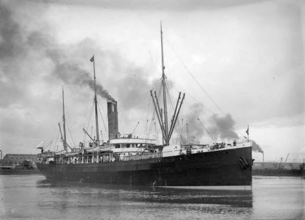 A vintage black and white photo of a large steamship with two smoke stacks releasing smoke. The ship sails on calm waters, with numerous masts and rigging visible. Flags are flying from its masts and stern. The background shows a cloudy sky.