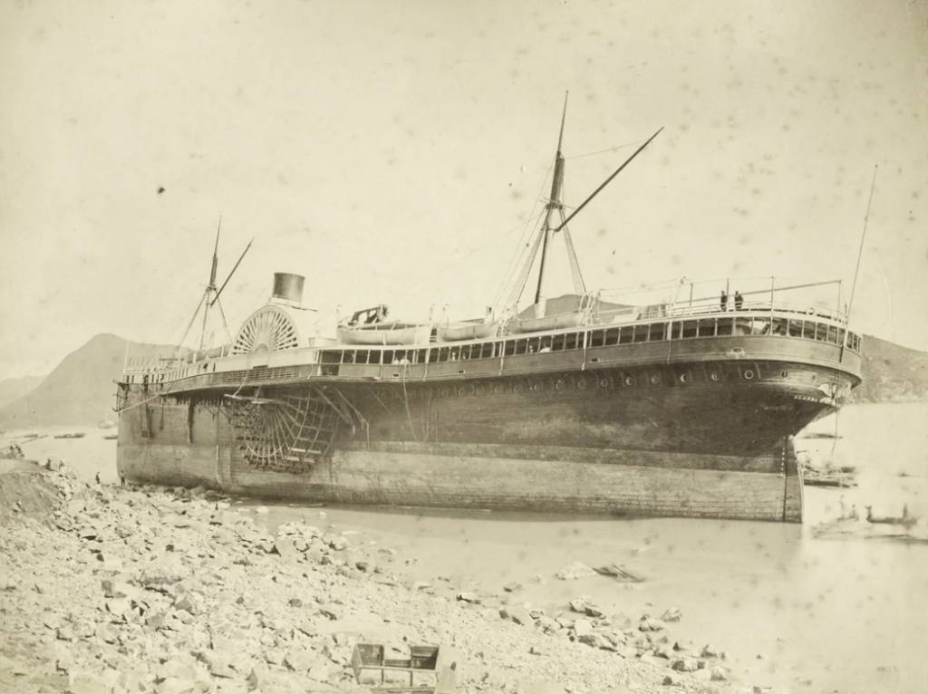 A vintage photograph of a large steamship tilted on its side near a rocky shore. The ship has visible paddle wheels and two tall masts. The surrounding landscape includes mountains in the background and calm water.