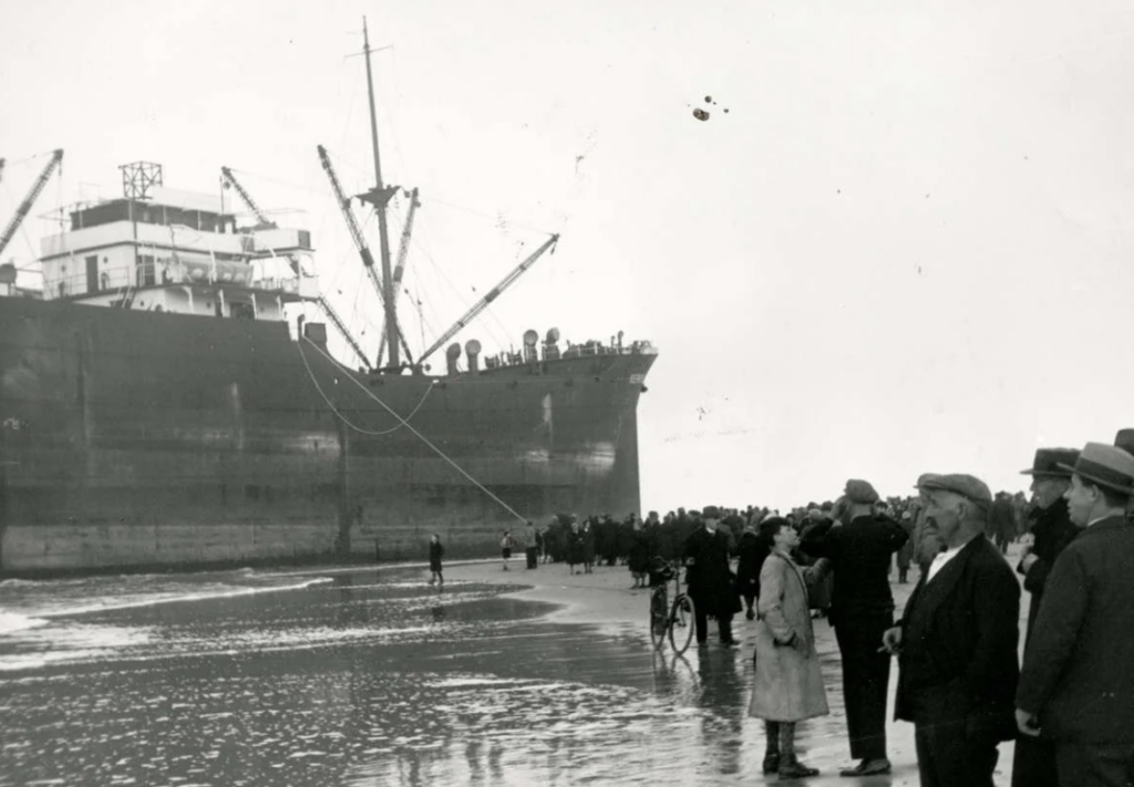 A large cargo ship is stranded on a beach, with people gathered around the shoreline observing. A line extends from the ship, and some people are closer to the water while others stand back, watching the scene.