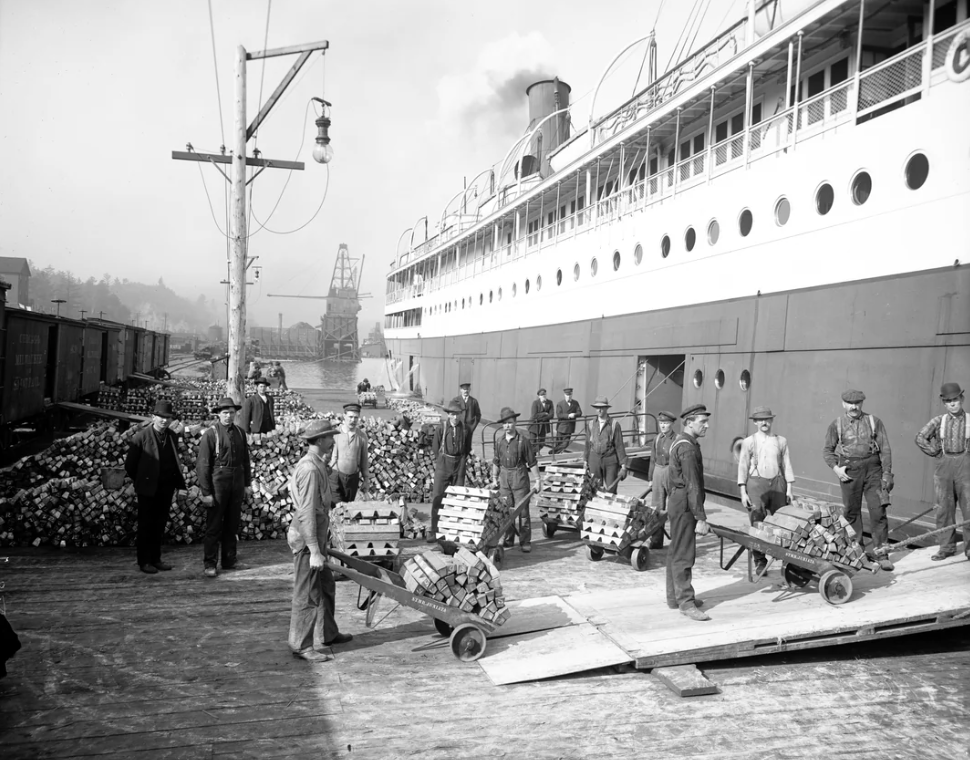 Workers load stacks of wood onto a large steamship docked at a pier. Men are using trolleys to transport the wood. The ship has a large funnel and multiple portholes. There is a crane and piles of wood in the background.