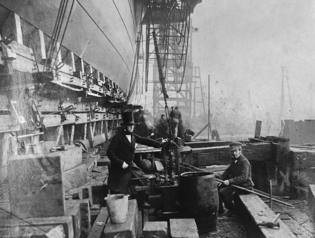 Historic black and white photo of shipbuilders at work. A man in a top hat stands beside machinery, while another sits on a barrel. Wooden beams, scaffolding, and the hull of a ship dominate the background.