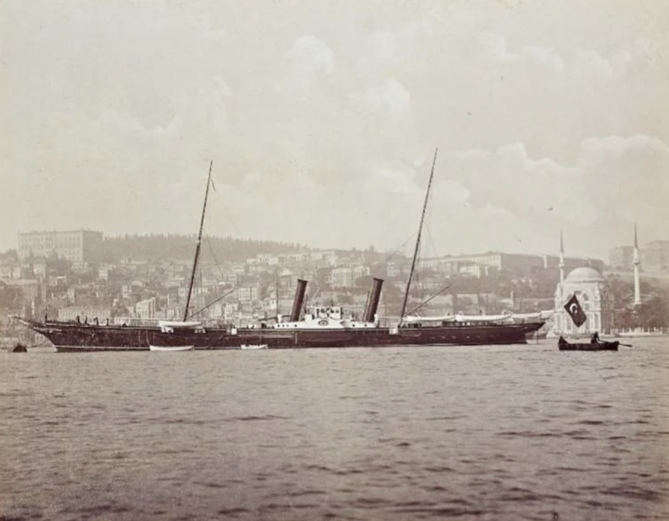 A vintage black-and-white photo of a steamship with two smokestacks on a calm body of water. In the background, a hilly cityscape with buildings is visible. A small boat with a large flag is in the foreground.