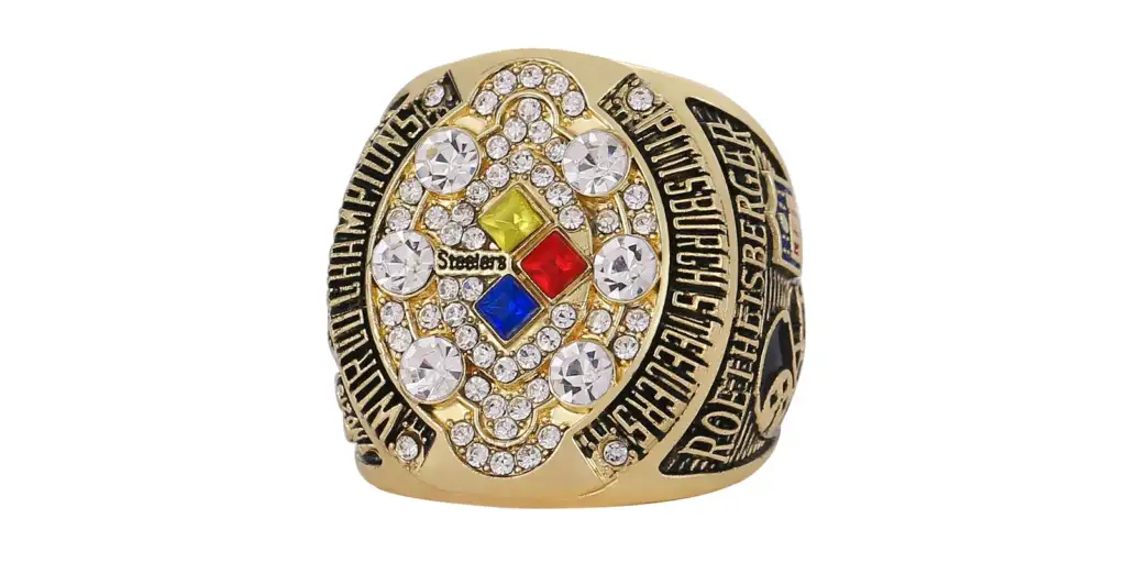 A gold championship ring with large diamonds forming an oval pattern in the center. The center square displays the word "Steelers" and colorful diamond shapes. Engravings and smaller diamonds cover the ring's surface, which signifies victory.