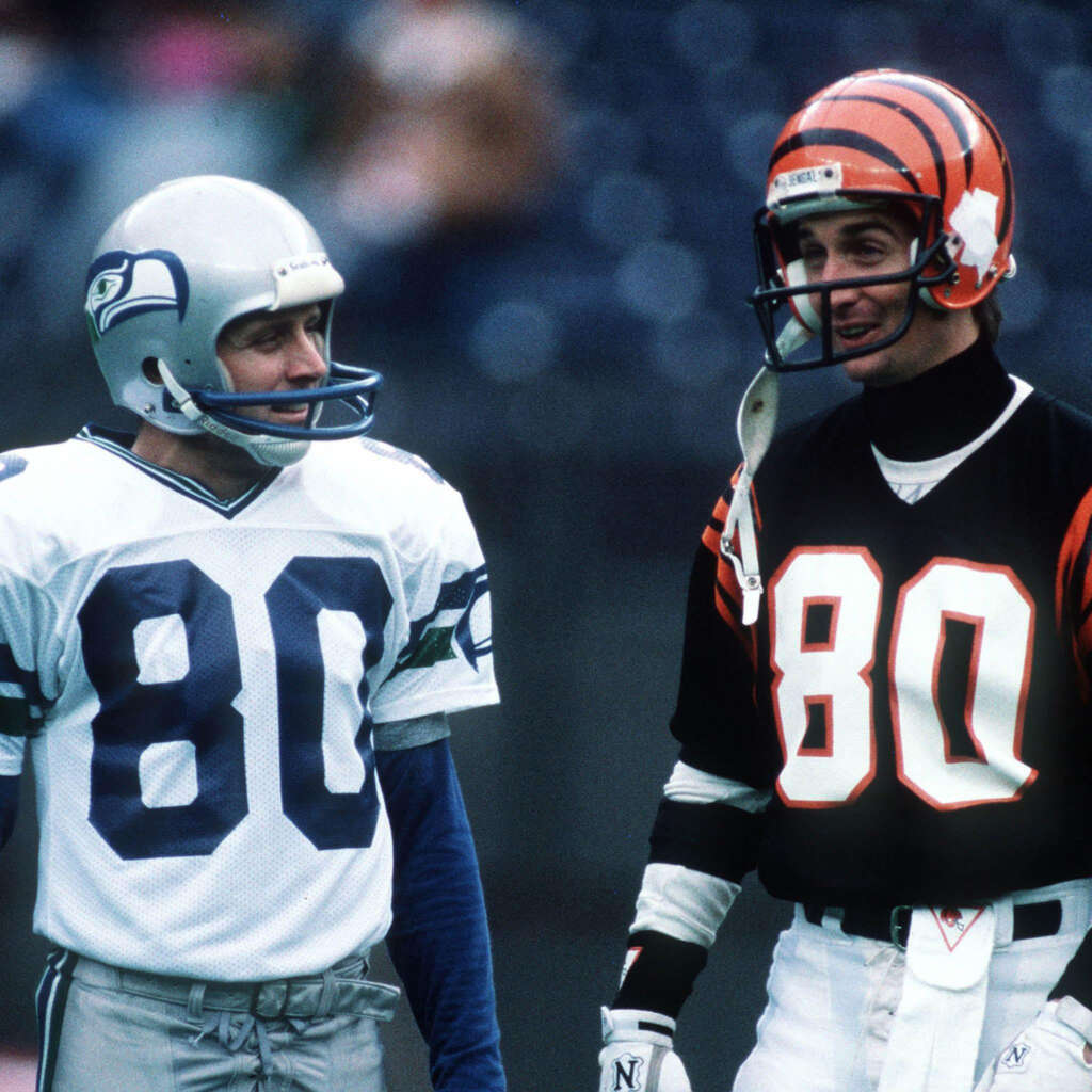 Two American football players stand on a field. One wears a white Seattle Seahawks uniform, and the other wears a black Cincinnati Bengals uniform. Both have helmets and jerseys numbered 80. They're engaged in conversation.