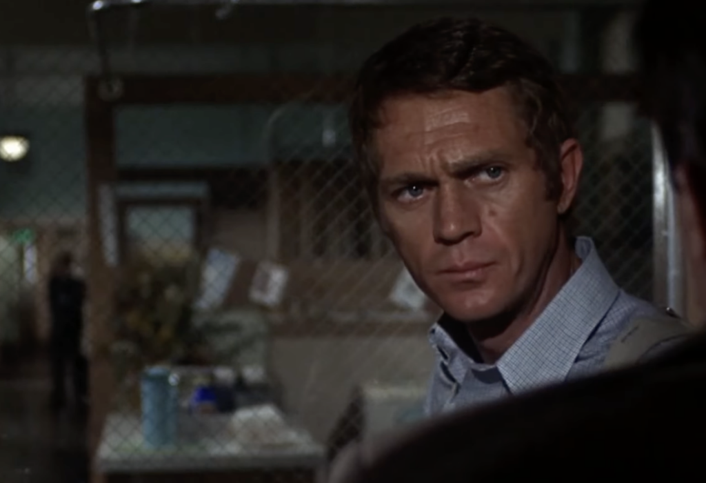 A man with short brown hair wearing a light blue shirt looks intently to his right in a dimly lit room with wire netting in the background. The setting appears to be an office with various items behind a mesh screen.