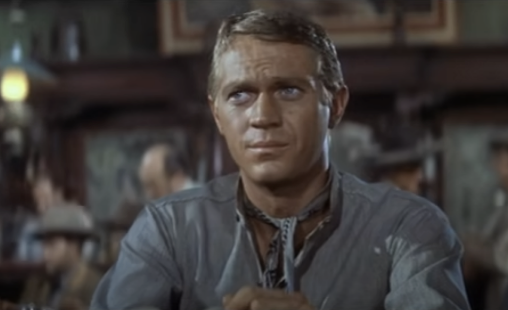 A man with short hair in a rugged shirt sits in a dimly lit bar or saloon, looking to the side with a serious expression. The background is blurred, showing people and dim lighting typical of a classic Western setting.