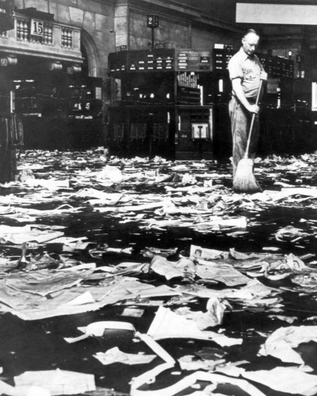 A man sweeps a floor covered in paper debris in a large room with high ceilings, appearing to be an office or financial space. Shelves and a clock are visible in the background, suggesting a busy environment.