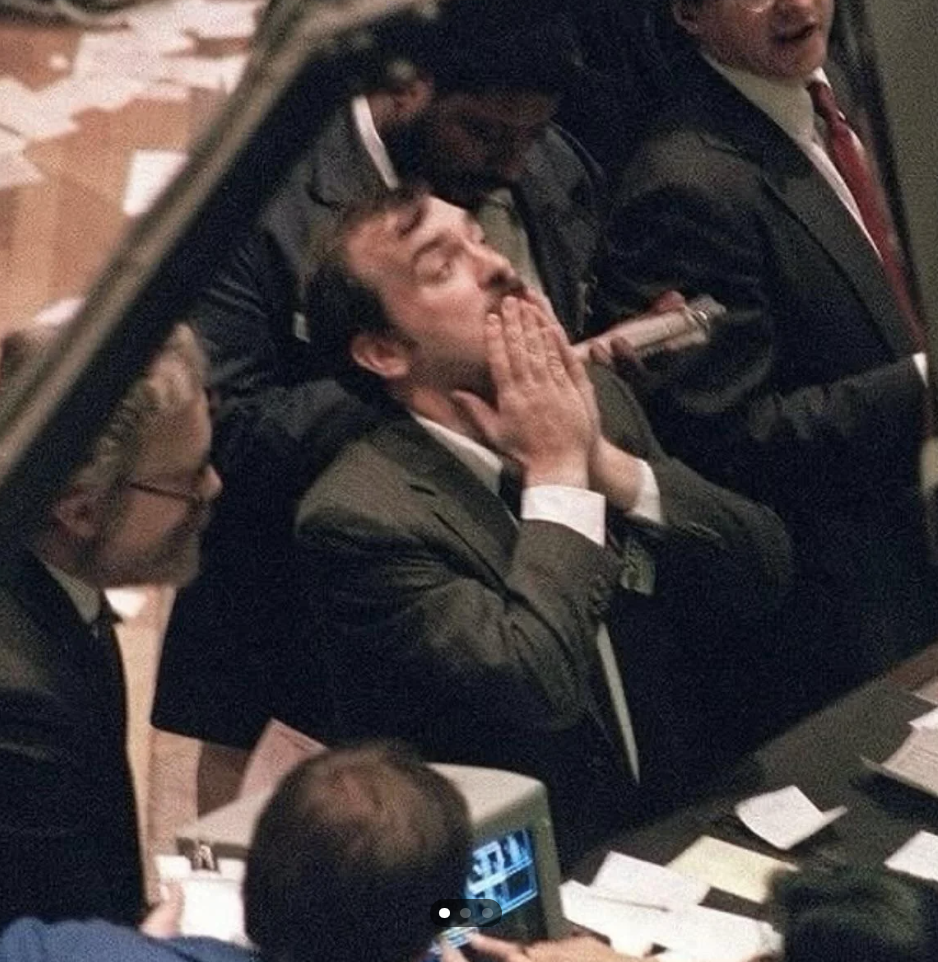 A group of people in formal attire gathered at what appears to be a busy trading floor. One man at the center has his hands over his mouth, looking concerned. Papers and monitors are visible around them, indicating a fast-paced environment.