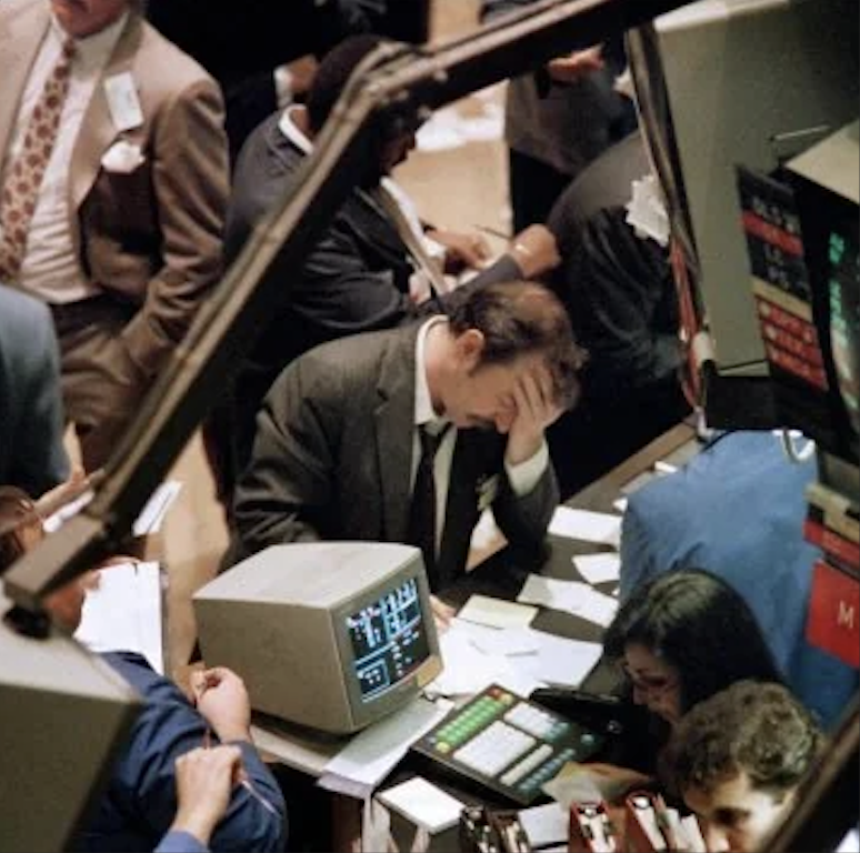 A man in a suit holds his head in his hand, appearing stressed, while seated at a cluttered desk with an old computer and papers. People stand around him in a busy office environment, suggesting a financial or trading setting.