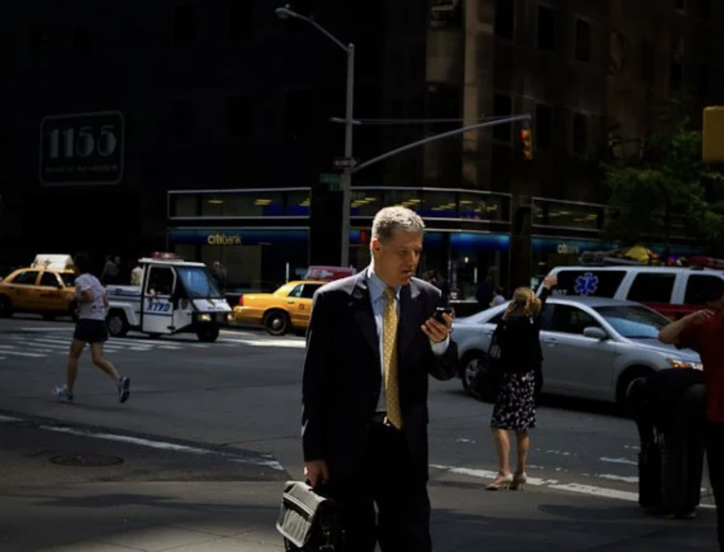 A man in a suit looks at his phone while standing on a busy city street. He's holding a briefcase. Taxis and pedestrians are in the background, with tall buildings visible. The scene is partially shaded.