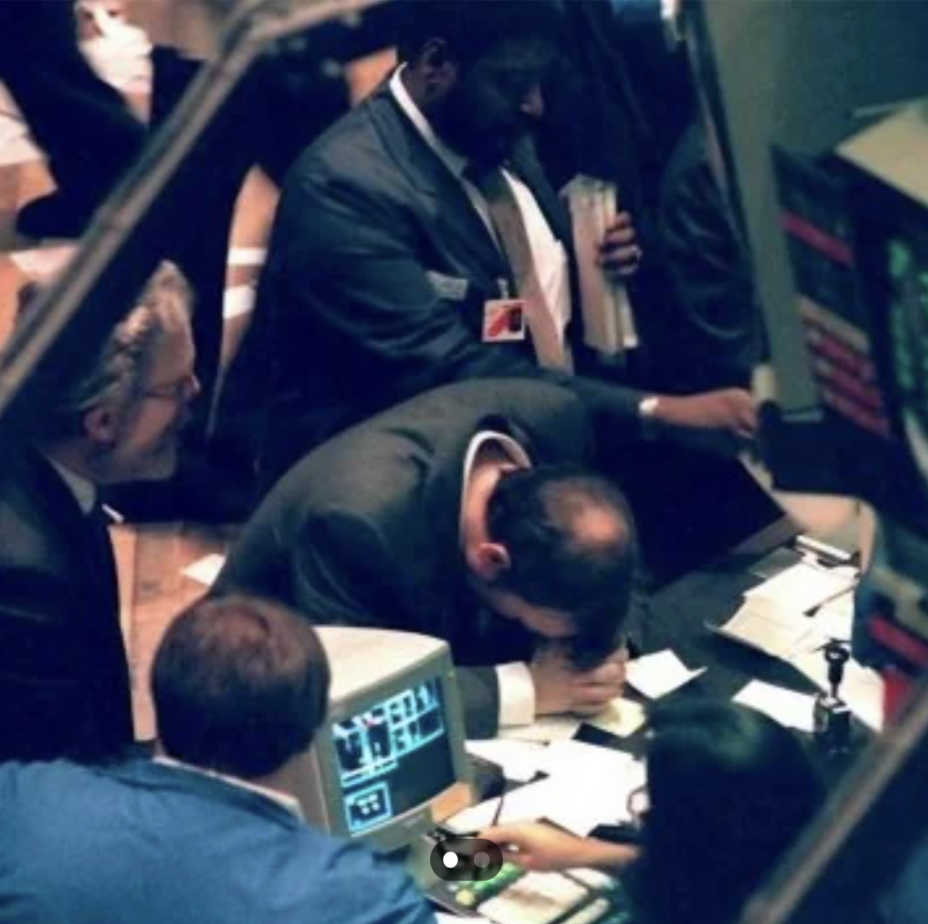 A group of people in business attire are gathered around a desk with computer monitors displaying data. One person sits with their head in their hands, suggesting stress or frustration. Others are standing and observing the monitors.