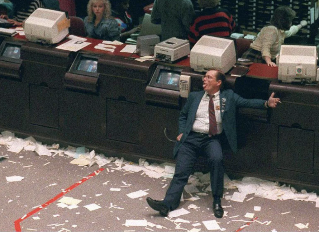 A man in a suit sits on the floor with papers scattered around, appearing exhausted or contemplative. He is leaning against a counter with computers and office equipment. People are visible in the background, seemingly busy at work.