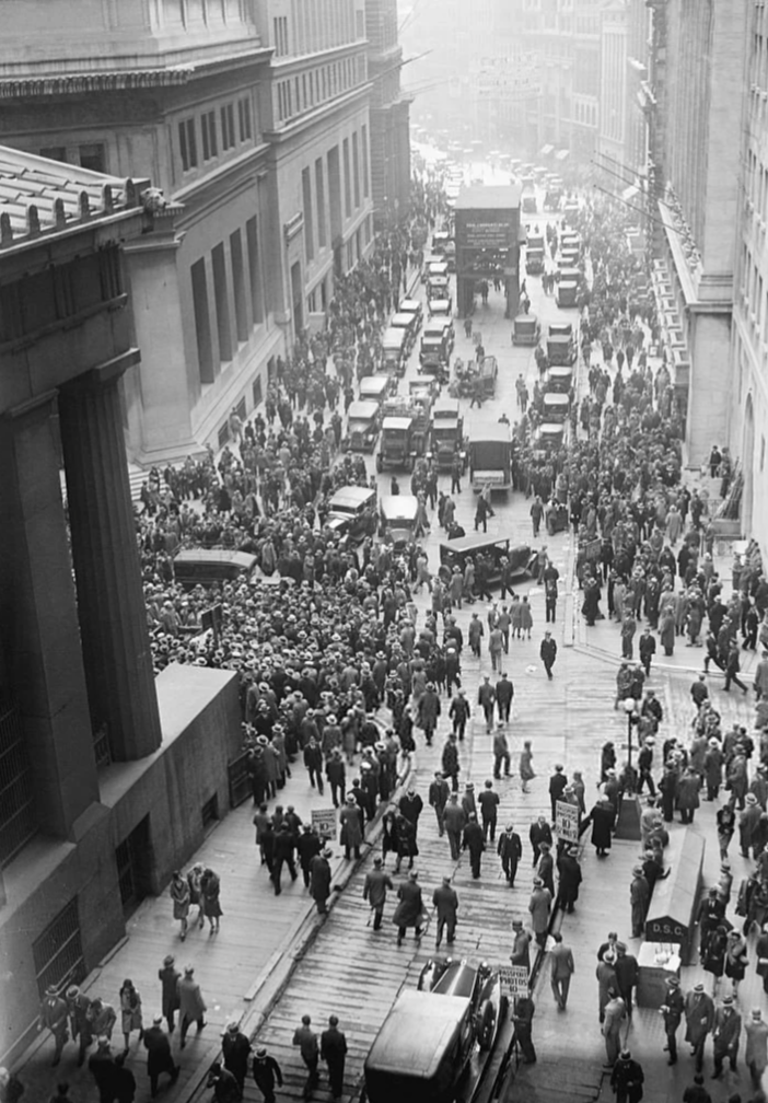 A black and white historical photograph depicts a crowded city street filled with people and vintage cars. The street is lined with tall buildings, and a large group is gathered around a central point in the road.