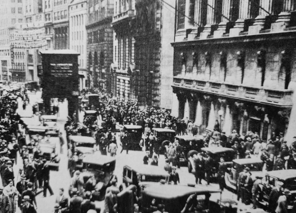 A black and white photograph of a busy early 20th-century urban street filled with people and vintage cars. Tall buildings line the street, and a large crowd is gathered, suggesting a bustling city scene.