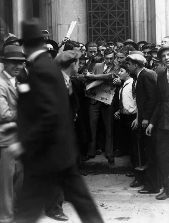 A group of men and boys gather on a city street, all focused on one person holding a newspaper. They are dressed in early 20th-century attire, with hats and suits. The scene is bustling with movement and energy.