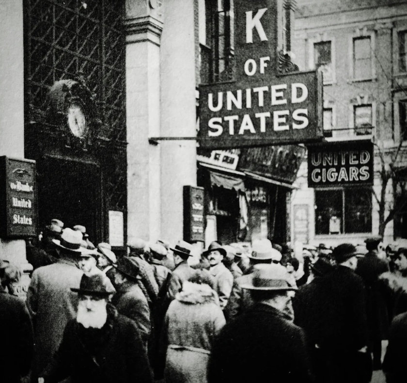 A black-and-white photo depicts a large group of people, mostly men in hats and coats, gathered outside the entrance of a United States bank. Signs reading "UNITED STATES" and "UNITED CIGARS" are visible on the building.