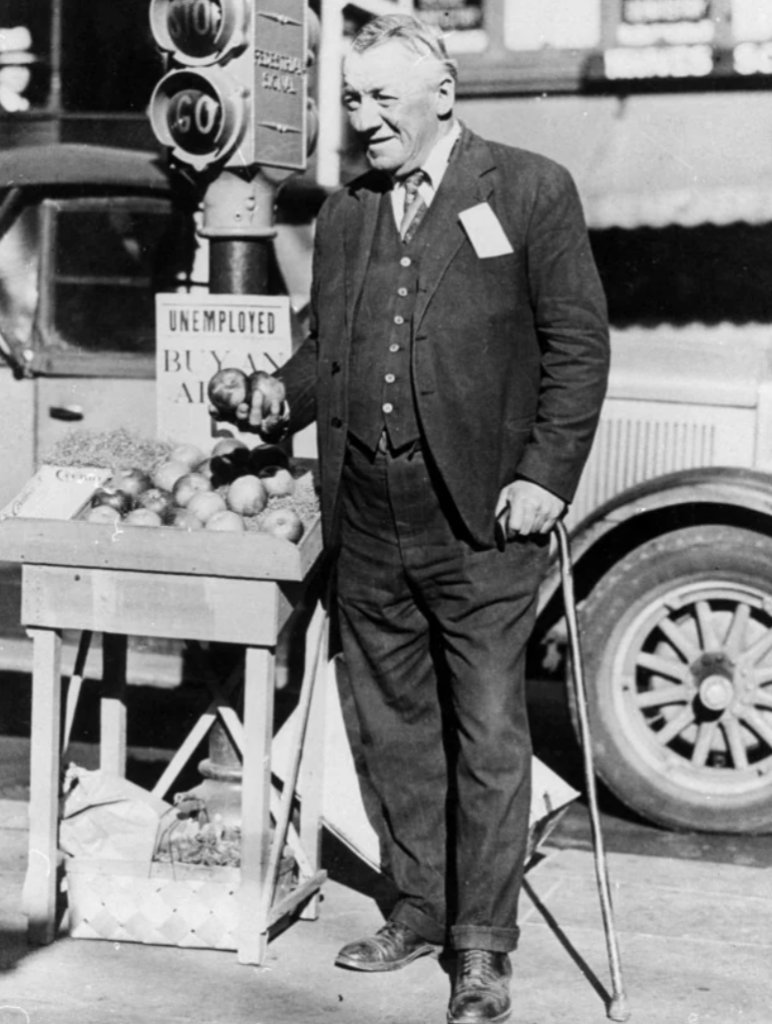 An elderly man with a cane stands next to a fruit stand labeled "Unemployed Buy Apples 5¢." He holds apples in one hand and is dressed in a suit. A vintage car and a traffic light are visible in the background. The scene appears to be from the early 20th century.