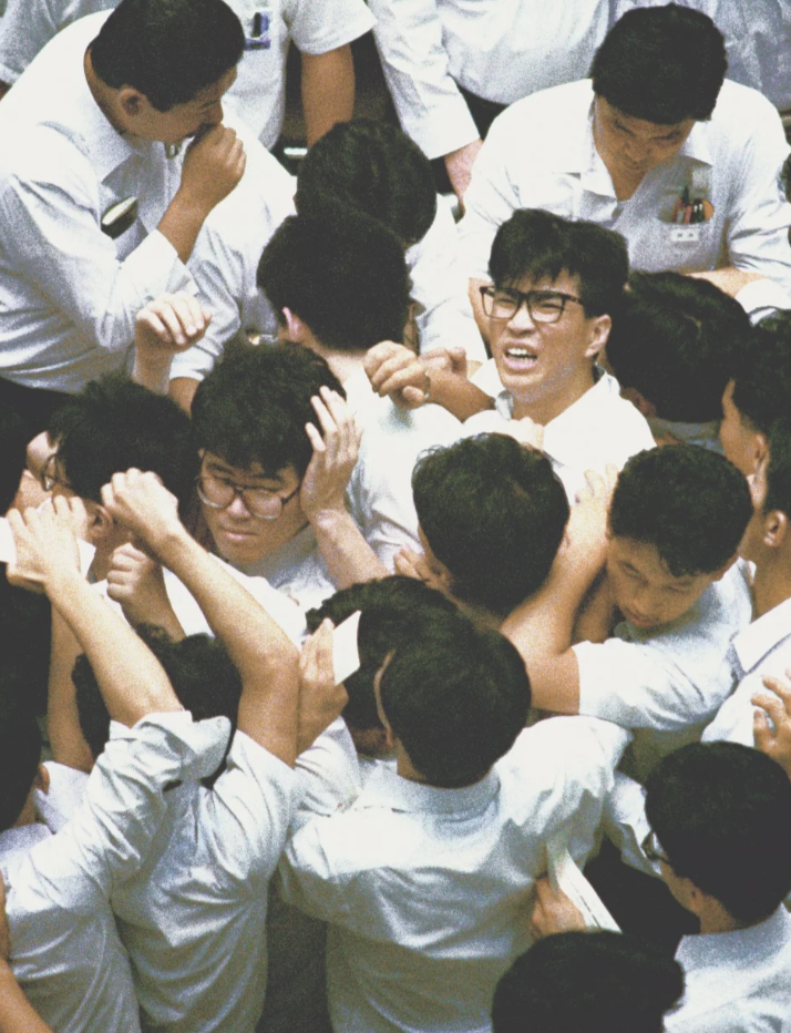 A large crowd of people in white shirts is tightly packed together. Some individuals are wearing glasses and appear to be engaged in a bustling, energetic exchange or activity, with expressions of excitement and determination.