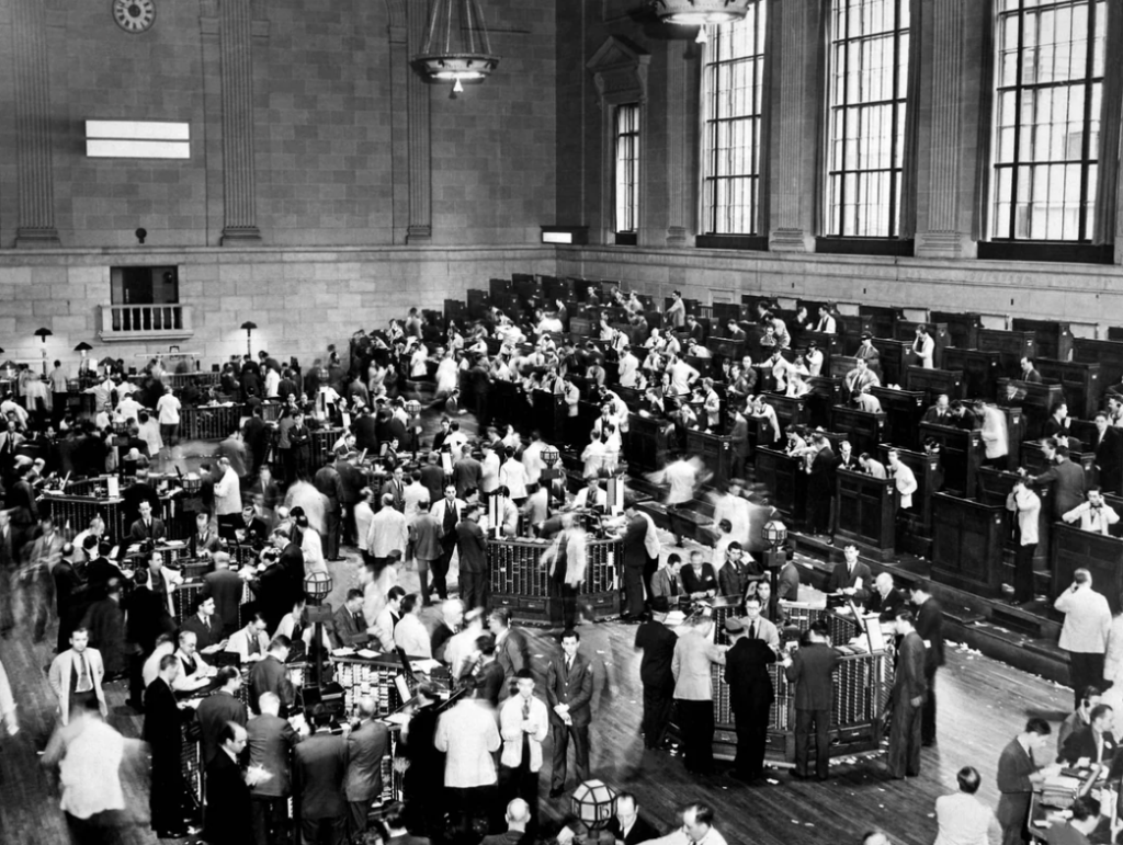 A historic black-and-white photo of a bustling stock exchange floor. People in suits are actively trading, gathered in groups, and surrounded by large wooden trading booths under tall windows, reflecting a lively financial atmosphere.