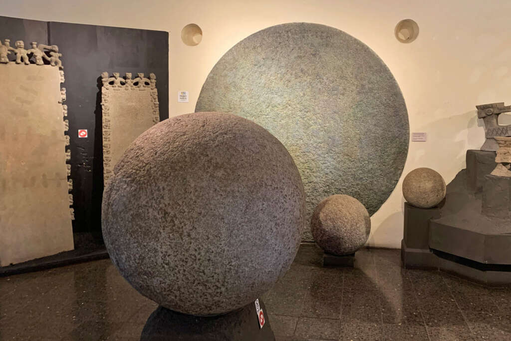 Several large, textured stone spheres are displayed in a museum room with smooth, dark flooring and neutral walls. Some spheres are freestanding, while others are mounted. The background includes stone carvings and subtle lighting.