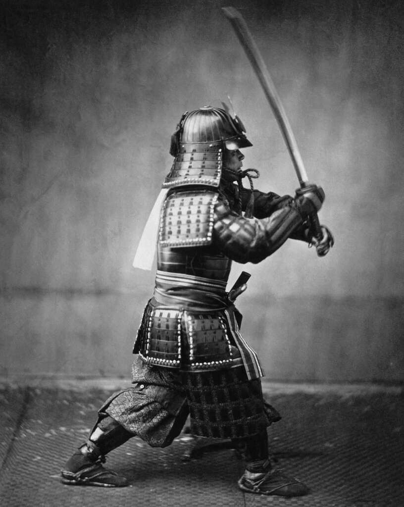 A historical black and white photo of a samurai in traditional armor, standing in a poised fighting stance. He holds a raised katana with both hands, ready for action. The background is plain, emphasizing his attire and posture.