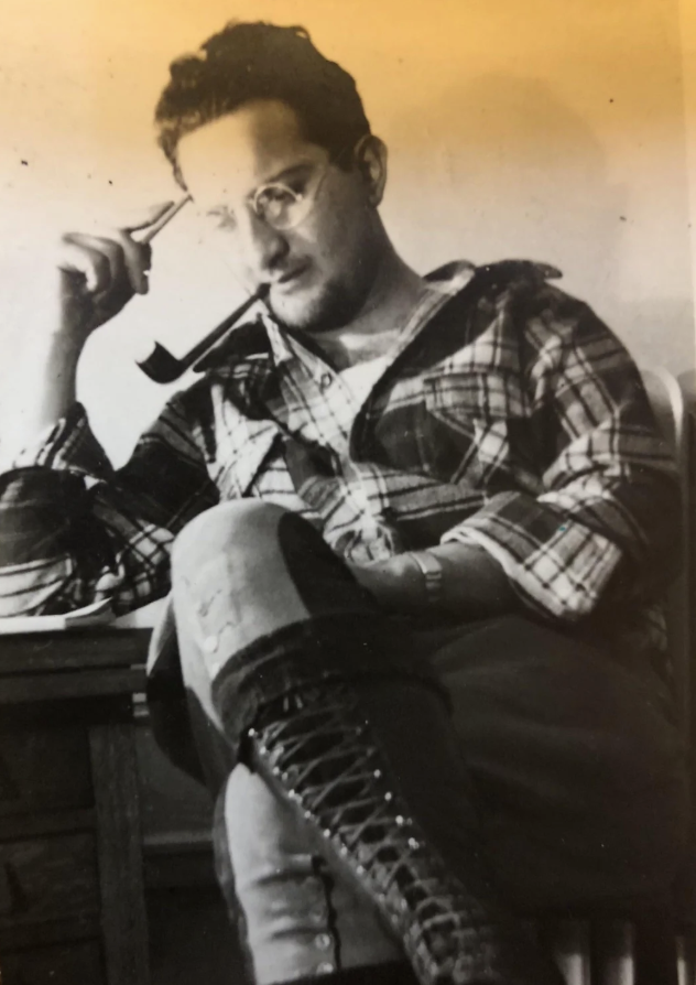 A black-and-white image of a man sitting, holding eyeglasses in one hand and a pipe in the other. He wears glasses, a plaid shirt, and laced boots. His legs are crossed, and he appears thoughtful, seated beside a table.