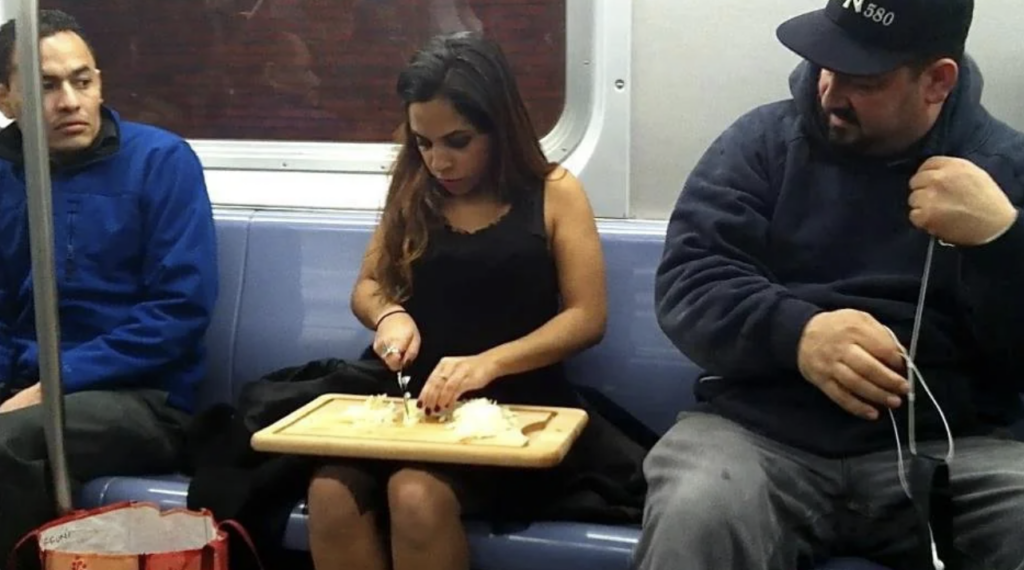 A woman sitting on a subway train cuts onions on a wooden chopping board placed on her lap. She is focused on her task while two men sit on either side; one looks away, and the other is wearing a cap, handling earphone wires.