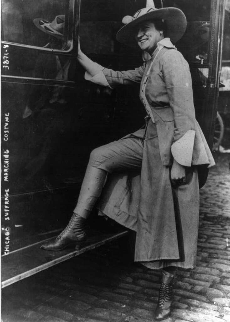 A woman in an early 20th-century outfit, wearing a wide-brimmed hat and a long coat, steps onto a platform. The image is labeled "Chicago suffrage marching costume." She stands on a cobblestone street, next to a vehicle.