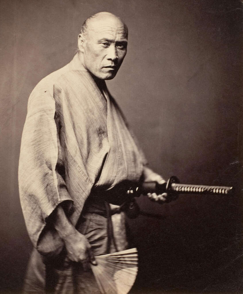 An older man in traditional Japanese attire poses with a katana in one hand and a fan in the other. His expression is serious and he stands against a plain background.