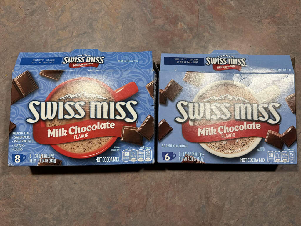 Two boxes of Swiss Miss Milk Chocolate hot cocoa mix are shown. The left box has eight servings, and the right has six. Both feature a red mug with cocoa and chocolate pieces on a blue background.