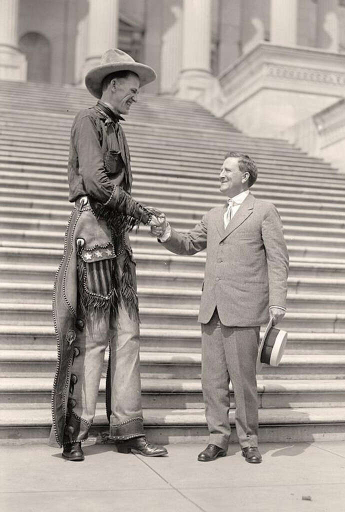 A very tall man in cowboy attire and a shorter man in a suit shake hands on the steps of a grand building. The cowboy wears a wide-brimmed hat and leather chaps, while the suited man holds a hat in his left hand.