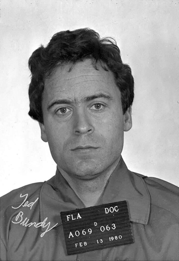 Black and white mugshot of a man with curly hair, wearing a shirt. He holds a plaque showing "Fla. DOC A069063" and the date "Feb 13 1980." The name "Ted Bundy" is handwritten in the lower left corner.