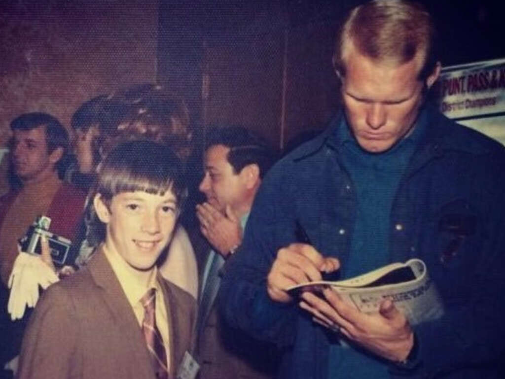 A young boy in a suit and tie smiles at the camera while a man beside him signs an autograph. The man is wearing a dark jacket and is focused on the paper in his hand. People and a poster board are visible in the background.